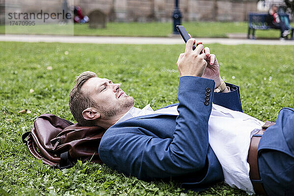Männlicher Berufstätiger benutzt Mobiltelefon  während er im Park auf Gras liegt