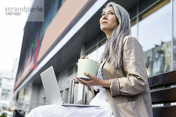 Nachdenkliche Frau hält eine Lunchbox in der Hand  während sie mit einem Laptop auf der Bank sitzt