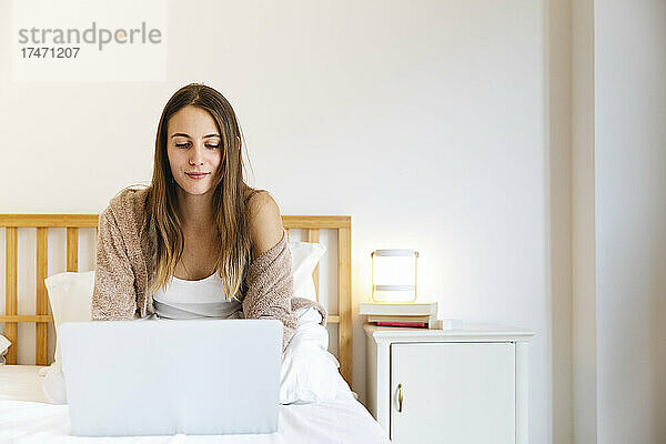 Junge Frau benutzt Laptop  während sie zu Hause im Bett sitzt
