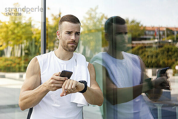 Sportler mit Mobiltelefon und Armbanduhr lehnt an Glaswand
