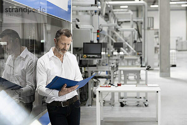 Reifer Geschäftsmann liest Akte  während er sich in der Fabrik an Glas lehnt