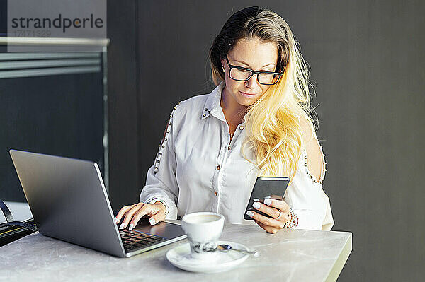 Blonde Geschäftsfrau benutzt Mobiltelefon  während sie im Café arbeitet