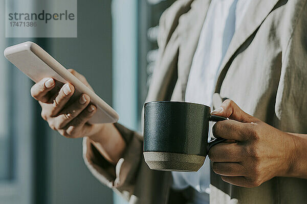 Frau hält Kaffeetasse in der Hand  während sie an einem sonnigen Tag ihr Mobiltelefon benutzt
