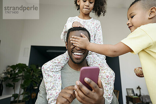 Verspielte Jungen und Mädchen haben Spaß mit dem Vater  der zu Hause sein Smartphone in der Hand hält