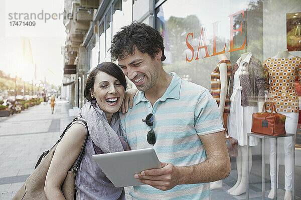 Fröhliches Paar nutzt digitales Tablet  während es neben dem Bekleidungsgeschäft steht