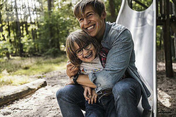 Vater umarmt Sohn  während er auf einer Rutsche im Wald sitzt