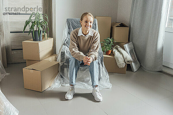Lächelnde junge Frau sitzt zu Hause auf einem in Plastik eingewickelten Sessel