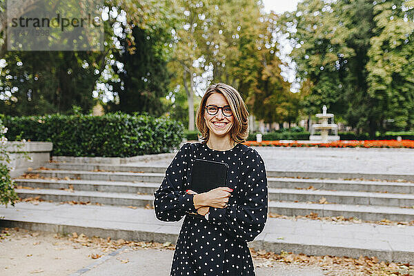 Lächelnde Frau hält digitales Tablet im öffentlichen Park
