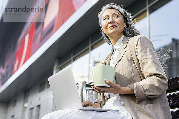 Reife Frau mit Lunchbox und Laptop sitzt auf Bank