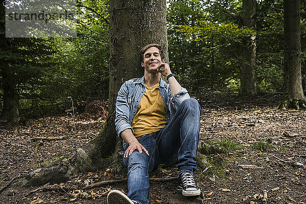 Lächelnder Mann mit geschlossenen Augen lehnt an Baumstamm im Wald