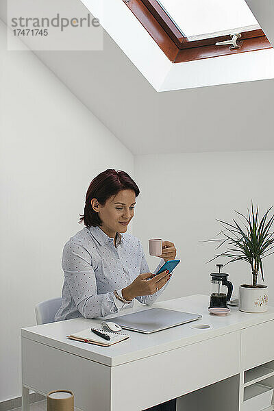 Geschäftsfrau nutzt Smartphone beim Kaffeetrinken am Schreibtisch im Heimbüro