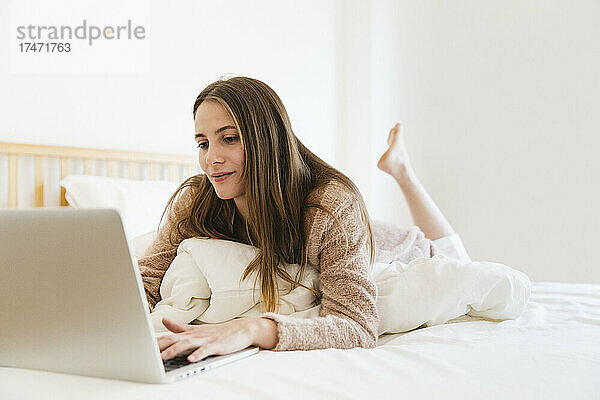 Junge Frau benutzt Laptop  während sie zu Hause im Bett liegt