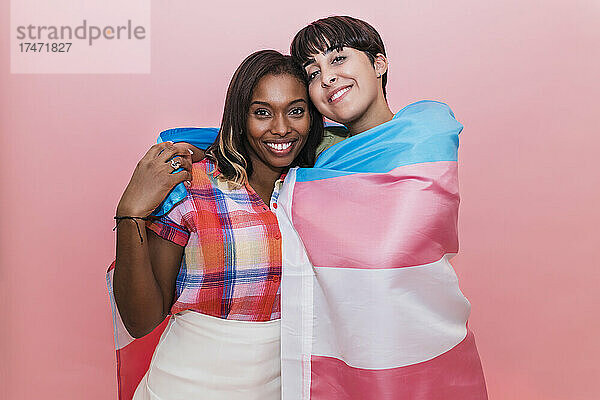 Lächelndes lesbisches Paar mit Transgender-Flagge vor rosa Hintergrund