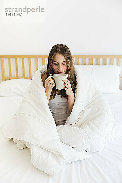 Junge Frau mit geschlossenen Augen trinkt Kaffee  während sie zu Hause in eine Decke gehüllt ist