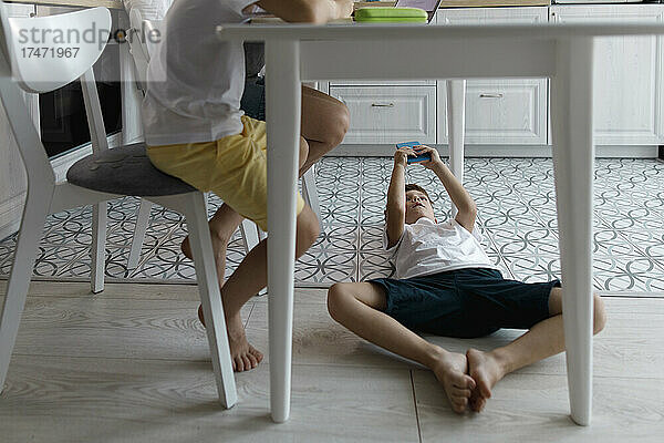 Junge benutzt Mobiltelefon unter Tisch  der auf dem Boden liegt