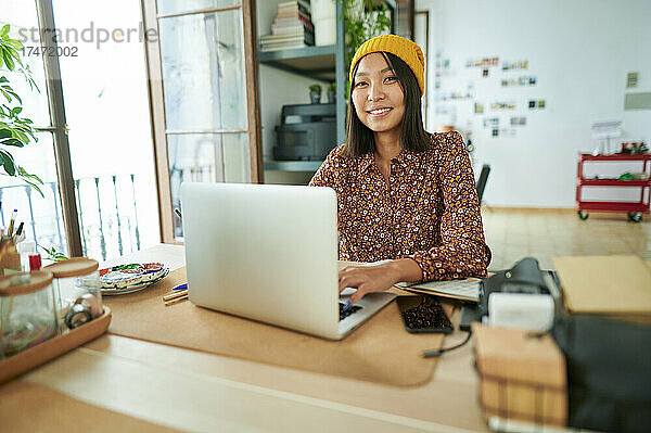 Berufstätige Frau mit Strickmütze sitzt mit Laptop am Arbeitsplatz