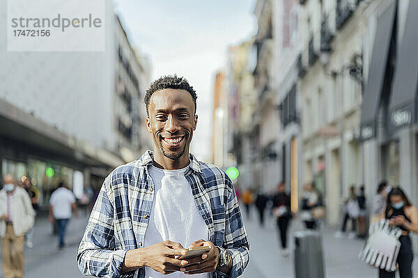 Lächelnder Mann im karierten Hemd mit Smartphone in der Stadt