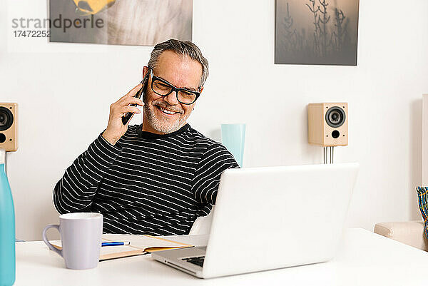 Glücklicher reifer Mann  der mit dem Smartphone spricht  während er im Heimbüro einen Laptop benutzt