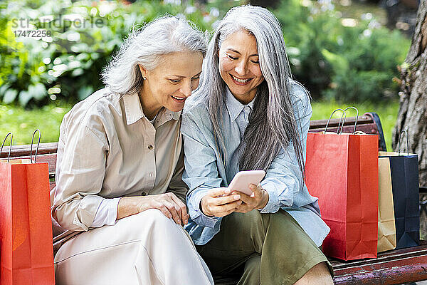 Reife Freundinnen mit Einkaufstüten teilen sich ihr Smartphone  während sie auf einer Bank sitzen