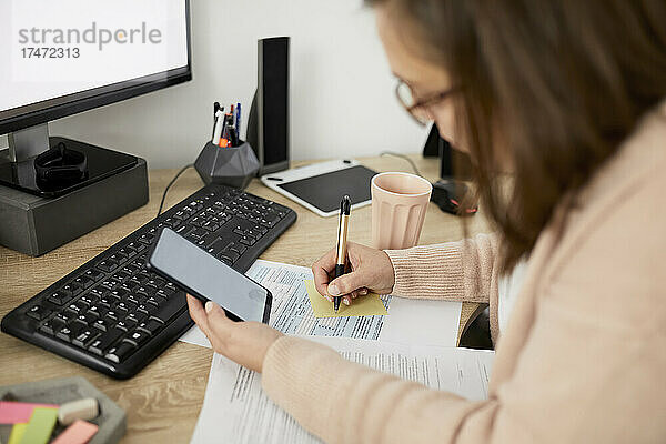 Geschäftsfrau hält Smartphone in der Hand und schreibt  während sie am Schreibtisch sitzt