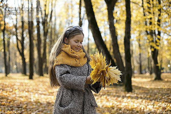 Lächelndes Mädchen mit Wollschal hält einen Haufen Herbstblätter im Park