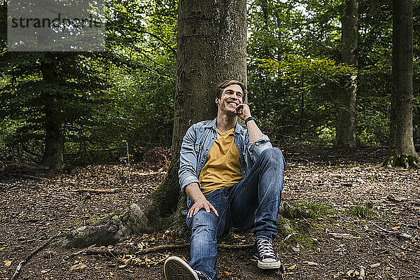 Glücklicher Mann mit der Hand am Kinn  der am Baumstamm im Wald sitzt