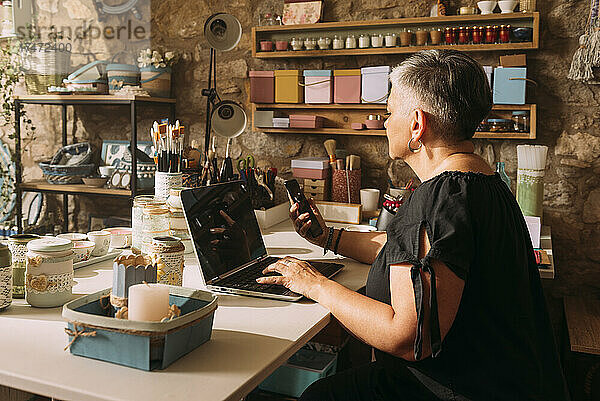 Reife Künstlerin mit Laptop und Mobiltelefon in der Werkstatt