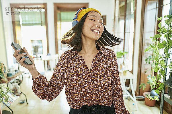 Lächelnde Geschäftsfrau wirft Haare  während sie im Büro Musik über Kopfhörer hört