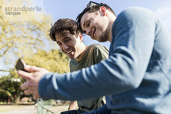 Lächelnde Männer  die im Park ihr Smartphone teilen