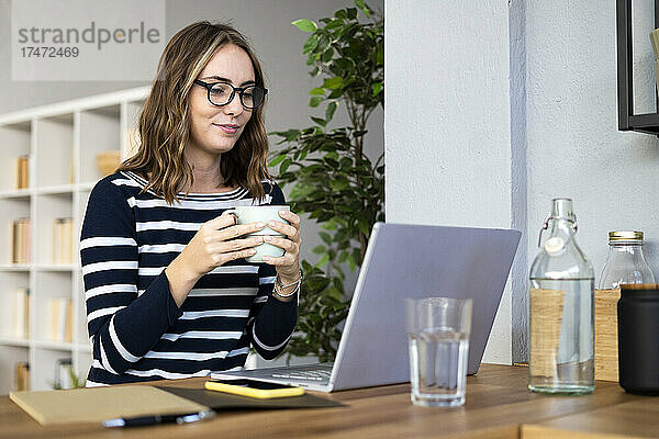 Lächelnde Frau trinkt Kaffee vor Laptop an der Küchentheke
