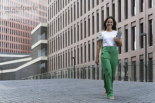 Lächelnde junge Geschäftsfrau geht mit Laptop auf Fußweg