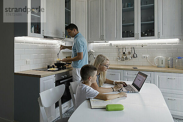 Mutter hilft Sohn bei den Hausaufgaben am Laptop am Tisch  während ein Mann in der Küche kocht