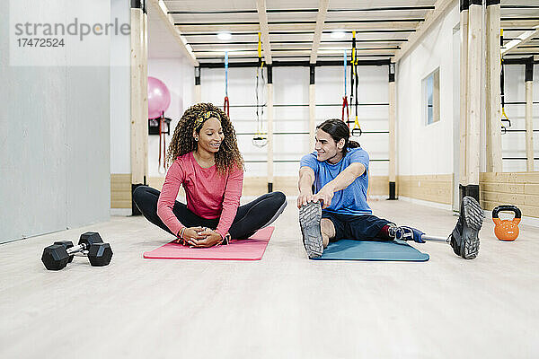 Lächelnde Frau macht Dehnübungen mit einer behinderten Freundin im Fitnessstudio
