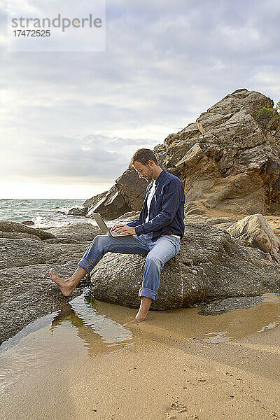 Mann sitzt auf einem Felsen und benutzt Laptop am Strand