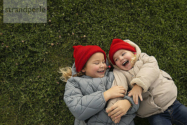 Girl in knit hat tickling sister while lying on grass in playground