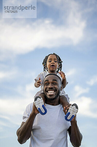 Fröhlicher Vater trägt Tochter auf Schultern unter dem Himmel