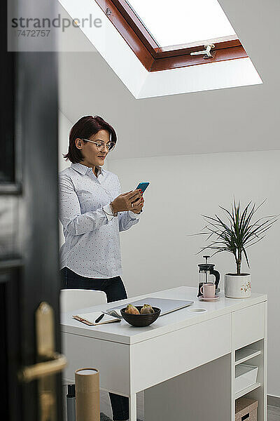 Geschäftsfrau schreibt Textnachrichten auf dem Smartphone  während sie im Heimbüro steht