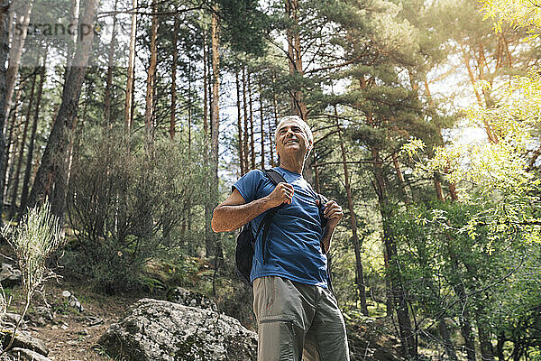 Lächelnder männlicher Tourist mit Rucksack  der im Urlaub im Wald steht