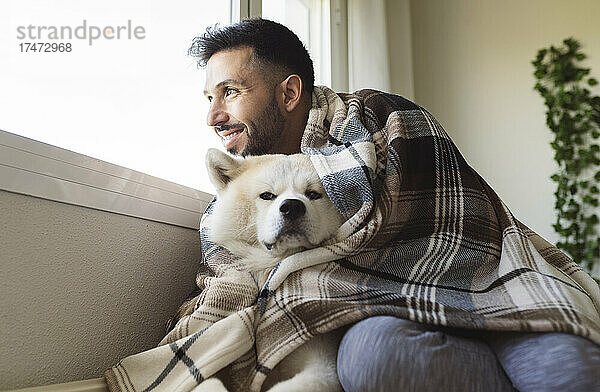 Lächelnder Mann mit Hund  der im Winter zu Hause durch das Fenster schaut