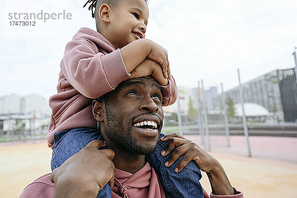 Lächelnder Vater schaut seinen Sohn an  der auf den Schultern im Sportplatz sitzt