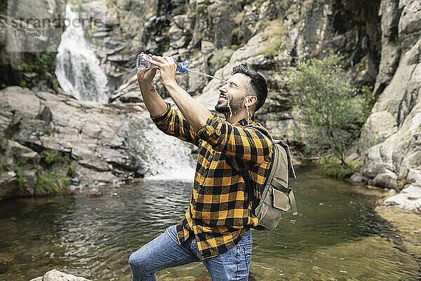 Lächelnder männlicher Tourist  der Wasser durch eine Flasche am Teich ins Gesicht schüttet