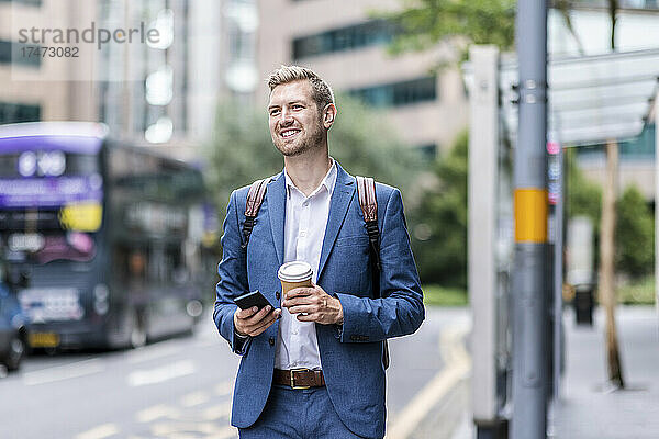 Lächelnder Geschäftsmann mit Einwegbecher und Mobiltelefon in der Stadt