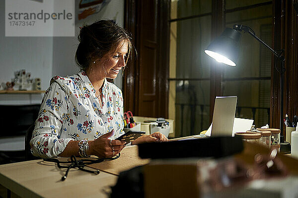 Geschäftsfrau hält Smartphone in der Hand  während sie im Büro am Laptop arbeitet