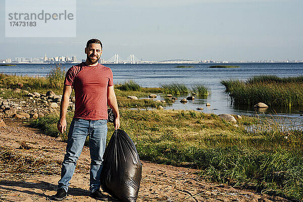 Lächelnder männlicher Umweltschützer mit Müllsack am Flussufer