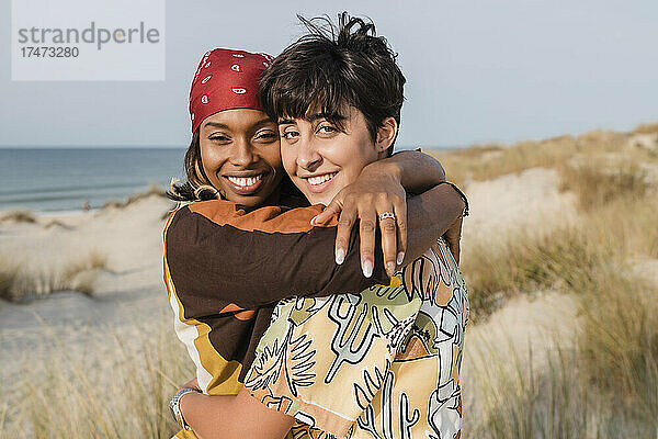 Lächelnde Frauen mit Kopftuch umarmen lesbische Freundin am Strand