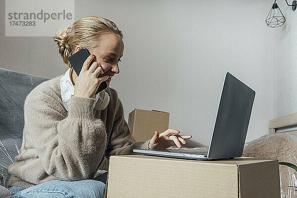 Lächelnde Frau mit Laptop  die im neuen Zuhause mit dem Handy telefoniert