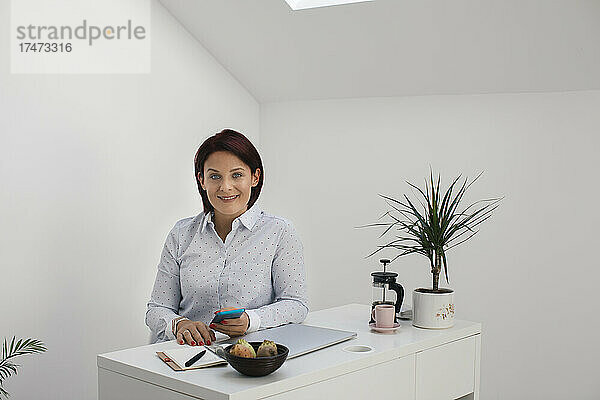 Lächelnder Freiberufler mit Smartphone vor der Wand im Heimbüro