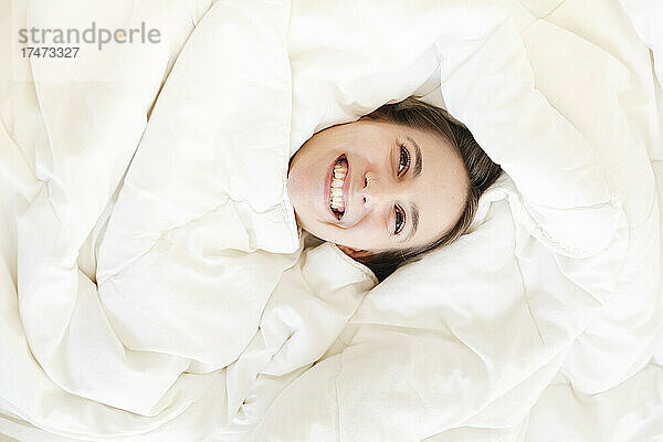 Glückliche junge Frau liegt mit weißer Decke im Schlafzimmer auf dem Bett