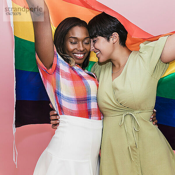 Glückliches lesbisches Paar mit Regenbogenfahne vor rosa Hintergrund