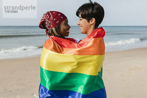 Glückliches lesbisches Paar mit Regenbogenfahne bedeckt  das am Strand steht